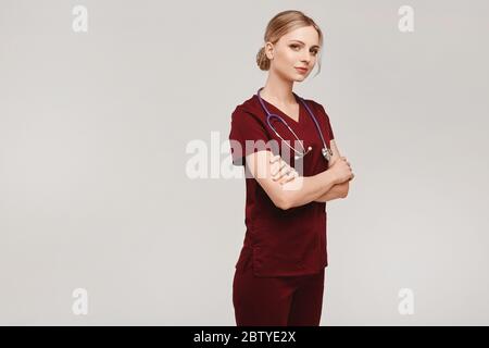 Jeune femme médecin avec stéthoscope posant avec des bras croisés sur le fond isolé. Jeune femme en uniforme médical posant sur fond gris Banque D'Images