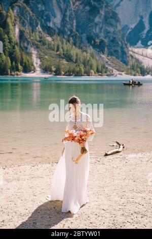 Belle mariée dans une robe blanche avec des manches et de la dentelle, avec un bouquet d'automne jaune de fleurs séchées et de roses de pivoine, sur le Lago di Braies en Italie Banque D'Images