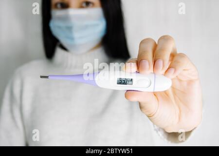 Jeune femme caucasienne en masque chirurgical tenant un thermomètre électronique dans les mains. Température élevée à l'écran, mise au point sélective Banque D'Images
