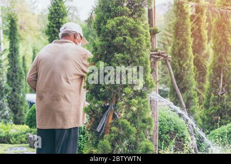 Un homme âgé en quarantaine porte un masque facial pour empêcher la propagation du virus Corona (Covid-19) arroser des plantes pour faire de l'exercice dans le jardin à la maison. Ne Banque D'Images