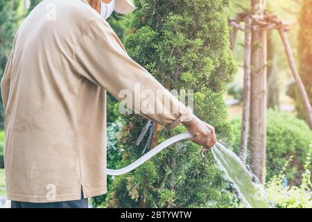 Un homme âgé en quarantaine porte un masque facial pour empêcher la propagation du virus Corona (Covid-19) arroser des plantes pour faire de l'exercice dans le jardin à la maison. Ne Banque D'Images