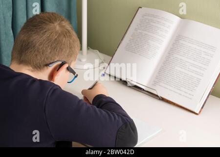 garçon dans des verres et une veste se trouve à une table et fait une tâche d'école Banque D'Images