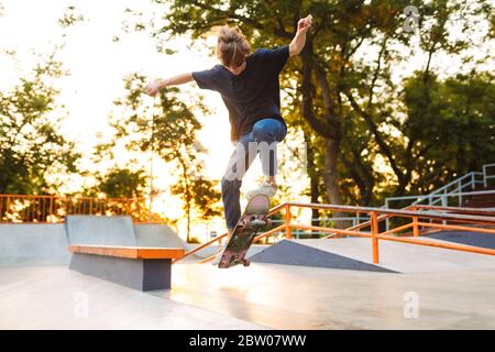 Jeune patineuse cool en T-shirt noir et jeans pratiquant des tours sur le skateboard au parc de skate moderne Banque D'Images