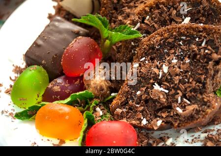 Morceaux de gâteau au chocolat et gelée colorée, bonbons au chocolat parsemés de flocons de noix de coco et chocolat sur une assiette blanche décorée de menthe près Banque D'Images