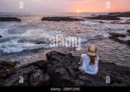Femme se trouve sur de vieilles roches de lave au bord de l'eau en regardant le coucher du soleil, Wawaloli Beach Park, Kailua-Kona, Hawaii, Etats-Unis. Banque D'Images