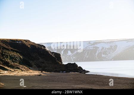 Une falaise au bord d'un lac couvert par la lumière du soleil le matin, avec la brume qui s'élève de la falaise, et de grandes montagnes en arrière-plan Banque D'Images