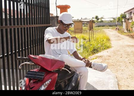 Un garçon indonésien à la peau brune habillé entièrement en blanc avec un chapeau, une montre et des bracelets sur son scooter rouge stationné dans une rue non pavée. En regardant Banque D'Images