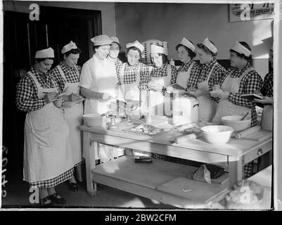 Les femmes sans emploi apprennent à être des cuisiniers cantines à Londres. Comme on s'attend à ce qu'il y ait bientôt une demande considérable de cuisiniers dans les institutions et les cantines, la Commission centrale pour la formation et l'emploi des femmes a ouvert deux centres de Londres où les femmes sans emploi entre 18 et 40 ans peuvent s'entraîner. Les femmes, qui doivent avoir perdu leur emploi à cause de la guerre, subiront un cours de six semaines durant lequel elles seront spécialement formées à la restauration et à la production de repas bon marché et nourrissants dans des conditions de temps de guerre pour un grand nombre. Les étudiantes se réunissent autour d'une table pour l'instruction Banque D'Images