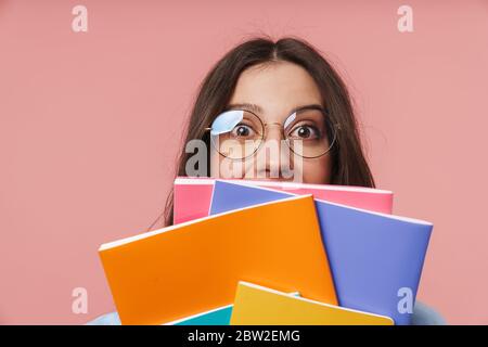 Image d'une jeune fille étudiante attrayante avec de longs cheveux bruns portant des lunettes tenant des livres d'exercice isolés sur fond rose Banque D'Images