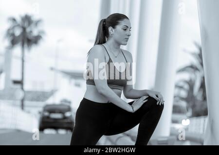 Jeune belle femme portant un soutien-gorge de sport et des collants étirant sa jambe gauche dehors en noir et blanc. Concept de mode de vie sain. Banque D'Images
