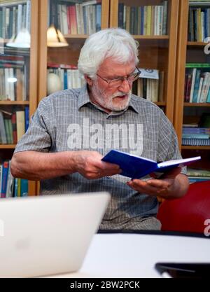 Portrait d'un homme âgé à barbe blanche avec chemise grise lisant un cahier bleu, livre en étant assis devant un immense étagère à livres en arrière-plan. Banque D'Images