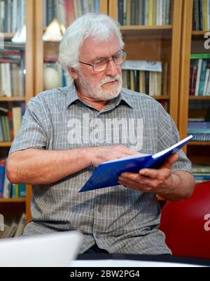 Portrait d'un homme âgé à barbe blanche avec chemise grise lisant un cahier bleu, livre en étant assis devant un immense étagère à livres en arrière-plan. Banque D'Images