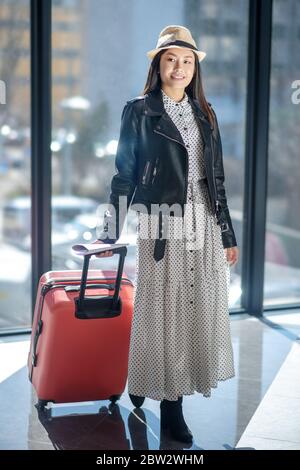 Brunette femme en cuir jacker et chapeau de paille marchant avec valise rouge, souriant Banque D'Images