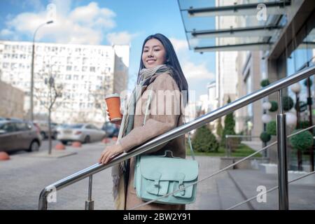 Brunette femme portant un manteau beige et un sac bleu, tenant une tasse de café, appréciant sa boisson, marchant dans les escaliers Banque D'Images