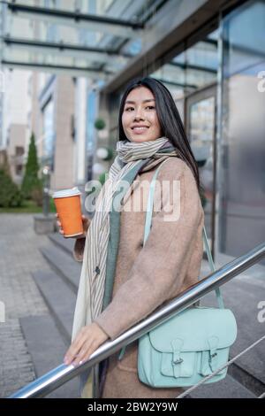 Brunette femme portant un manteau beige et un sac bleu, tenant une tasse de café, appréciant sa boisson, marchant dans les escaliers, souriant Banque D'Images