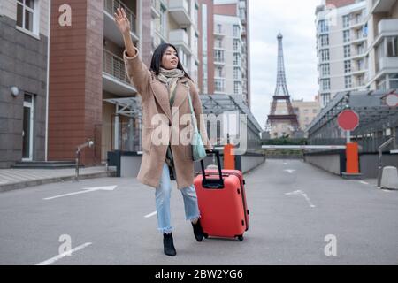 Brunette femme en beige manteau marchant avec une valise rouge, essayant de prendre un taxi Banque D'Images