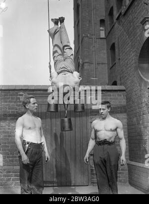 Cirque acrobat hommes forts pratiquant une routine dans une cour en brique . Un homme d'acrobat est suspendu à des anneaux avec des poids sur son cou et ses mains tandis que deux autres regardent dessus Banque D'Images