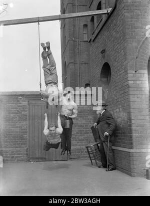 Cirque acrobat hommes forts pratiquant une routine dans une cour en brique . Trois hommes ont équilibré sur des anneaux de suspension tandis qu'un homme portant une chaise et une canne regarde dessus Banque D'Images