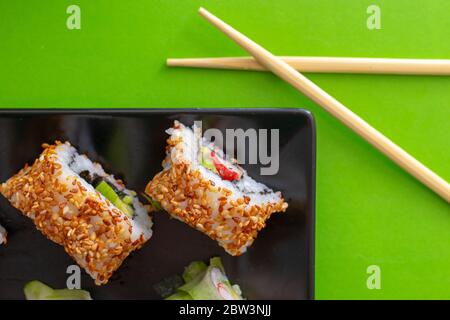 Vue de dessus d'une assiette carrée noire avec plusieurs petits pains à sushis aux graines de sésame et au concombre sur fond vert simple avec baguettes croisées Banque D'Images