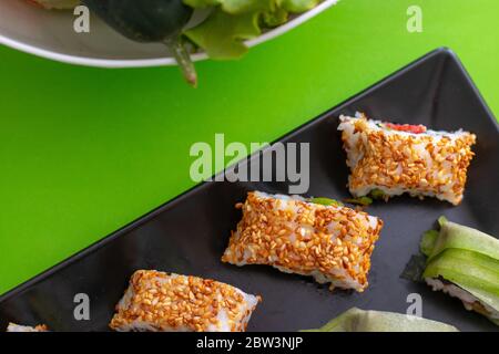 Vue de haut en bas d'une assiette de sushis noirs avec des rouleaux de californie et de concombre avec des graines de sésame. Fond vert simple et un bol de légumes Banque D'Images