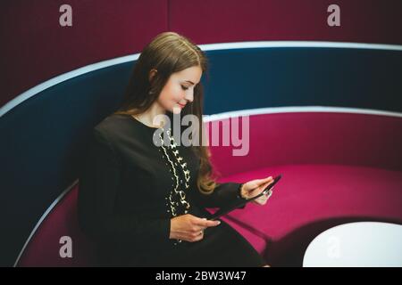 Une charmante jeune femme d'entrepreneur hispanique est assise sur un canapé rond lilas et analyse de la lecture sur l'écran d'une tablette numérique entre ses mains Banque D'Images