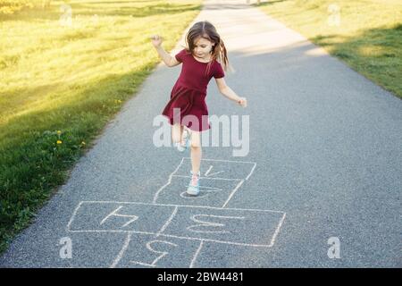 Mignon adorable petite jeune fille chld jouant le hopscotch en plein air. Jeu d'activités amusant pour les enfants sur l'aire de jeux à l'extérieur. Sports de rue de cour d'été Banque D'Images