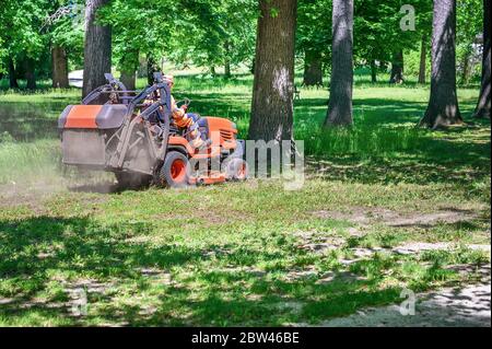 L'homme tond l'herbe sous les arbres dans le parc en faisant rouler la machine de tonte par temps ensoleillé Banque D'Images