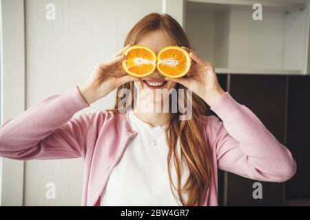 Rouge cheveux caucasienne femme souriant à la caméra tout en couvrant ses yeux avec des oranges dans la cuisine Banque D'Images