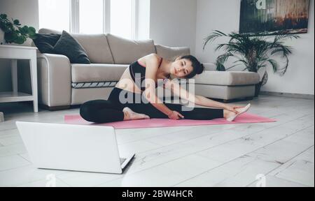 Brunette dame dans les vêtements de sport s'étirant sur le sol regardant l'ordinateur portable et faisant des exercices de yoga Banque D'Images