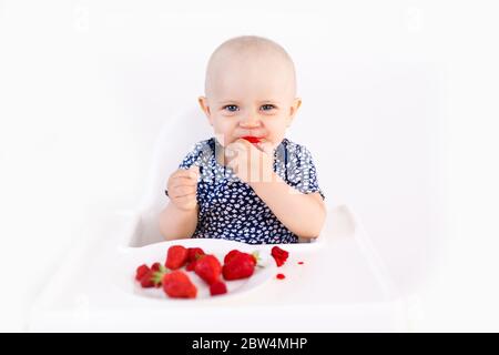 Bébé fille assis dans la chaise haute d'enfant manger des baies sur un fond blanc. Concept de nourriture pour bébé, espace pour le texte Banque D'Images