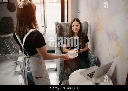 Une femme Barista tient un latte à une fille dans un café. Une femme qui a de longs cheveux travaillant à distance sur un ordinateur portable garde les distances sociales saisir une tasse o Banque D'Images