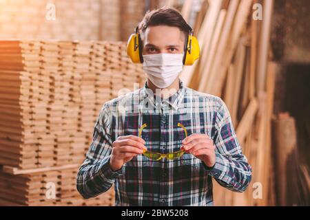 Portrait de menuisier professionnel portant un masque médical, des écouteurs de protection auditive, des lunettes de protection à la scierie. Artisan masculin en protecti Banque D'Images