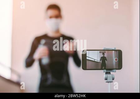 Jeune homme de sport blogger influenceur dans le masque de protection enregistrement vidéo tutoriel pour l'entraînement à la maison. Homme sportif confiant dans un masque médical Banque D'Images