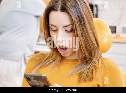 Patiente surprise assise sur une chaise et lisant des nouvelles choquantes sur un smartphone lors d'une visite chez un dentiste en clinique Banque D'Images