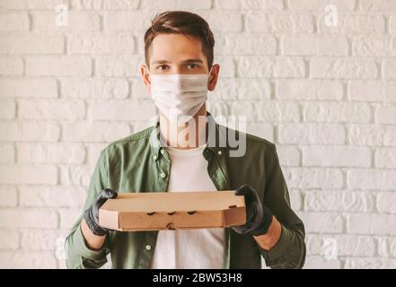 Bon livreur dans un masque médical et des gants tenant une boîte à pizza en carton dans les mains. Hipster man italien employé de service de livraison rapide de nourriture dans pr Banque D'Images