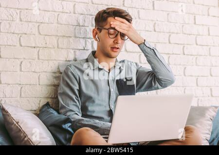 Un jeune homme hipster stressé en lunettes se sent fatigué, épuisé pendant que l'ordinateur portable est utilisé sur le canapé. Un homme d'affaires freelance et nerveux semble malheureux ou dépreux Banque D'Images