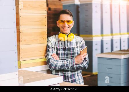 Heureux samwill propriétaire d'affaires dans des lunettes de protection et des écouteurs souriant avec des bras croisés tout en inspectant l'inventaire de stockage. Gestion d'entrepôt jeune Banque D'Images