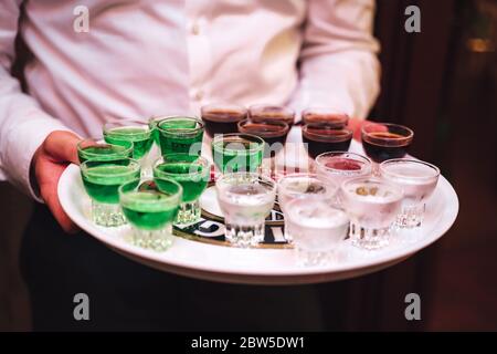 Homme en chemise portant un plateau avec plusieurs boissons alcoolisées. Serveur servant des tirs. Célébration, anniversaire, fête, mariage. Banque D'Images