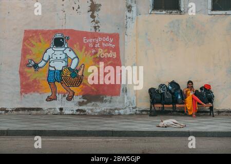 Une femme attend avec ses bagages sur un banc à côté de l'art de rue avec le texte "tout le monde est à la maison à Pondy" à Pondichéry, Inde Banque D'Images