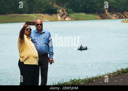 Deux hommes plus âgés prennent un selfie à Echo point, sur le réservoir du barrage Mattupatti à Munnar, Kerala, Inde. Un bateau de touristes passe derrière Banque D'Images