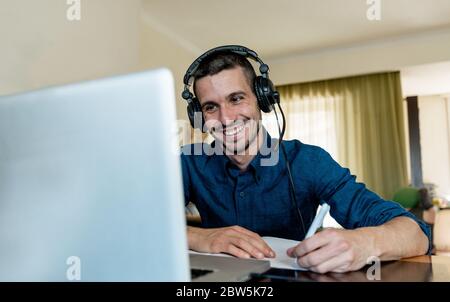 Un jeune étudiant heureux à la maison étudiant sur un ordinateur portable portant un casque écrivant sur un livre avec un stylo. Concept de cours en ligne. Banque D'Images