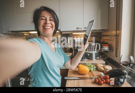 Femme âgée prenant un selfie cuisant des légumes avec l'aide de recette sur tablette dans la cuisine à la maison Banque D'Images
