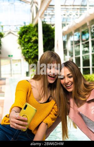 Deux jeunes filles caucasiennes attrayantes aux cheveux blonds sont photographiées avec leur téléphone mobile dans une fontaine à l'intérieur d'un bâtiment blanc lumineux Banque D'Images