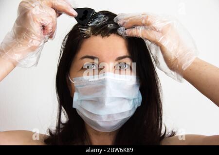 La femme brune, dans un masque médical et des gants à la maison, teint ses cheveux elle-même en quarantaine, Covid-19 Banque D'Images