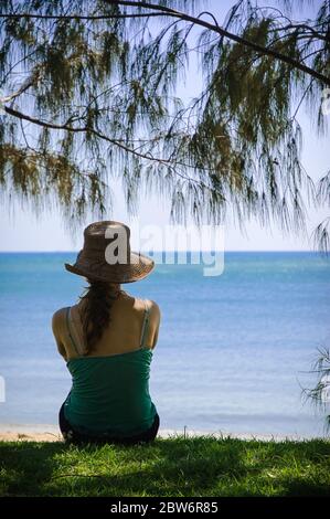 Assise touristique féminine, encadrée sous une Casuarina, sur une plage gazée donnant sur l'océan Pacifique à Palm Cove, à l'extrême nord du Queensland. Banque D'Images