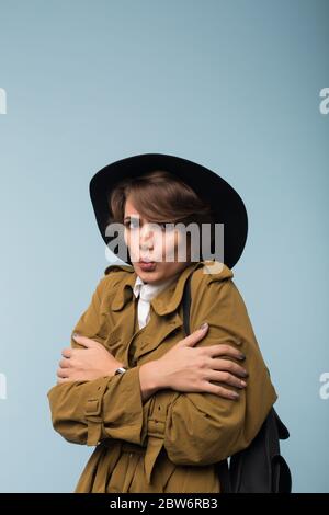 Jeune femme bouleversée avec des cheveux courts sombres dans un trench coat et un chapeau regardant malheureusement dans l'appareil photo tout en secouant du froid sur fond bleu Banque D'Images