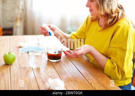 Vue latérale de la récolte femelle d'âge moyen prenant remède de bouteille en plastique tout en étant assis à la table avec une tasse de thé et un masque de protection à la maison Banque D'Images