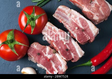 Tranches de porc crues sur un tableau d'ardoise noire, tomates, poivron rouge et poudre. Ventre de porc avec légumes. Photo de haute qualité Banque D'Images