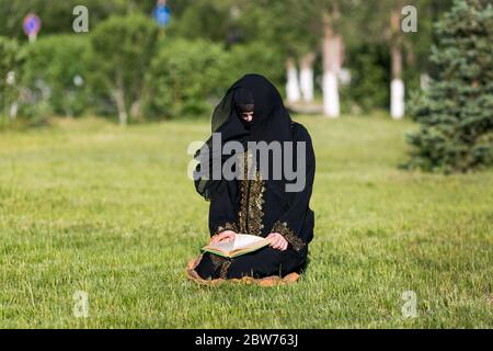 La femme islamique effectue la prière du matin, assise sur de l'herbe verte dans un parc Banque D'Images