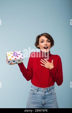 Jolie fille surprise avec cheveux courts foncés dans le chandail rouge étonnement regardant dans l'appareil photo avec peu de boîte présente dans la main sur fond bleu isolé Banque D'Images